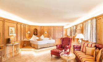 A cozy hotel room with wooden walls and elegant furniture. There is a comfortable bed, a seating area with a red armchair, and warm lighting.