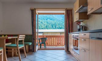 A cozy kitchen with wooden cabinets and a dining table. From the open door, there is a beautiful view of the mountains and nature.