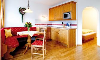 A cozy kitchen with a red dining area and wooden shelves. The decoration includes flowers and a warm atmosphere.