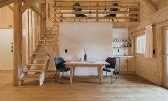 A cozy wooden apartment with an open kitchen and a dining area. The stairs lead to a small balcony on the upper floor.