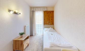 A simple room with a single bed and a wooden wardrobe. Natural light streams through the curtains, creating a bright atmosphere.