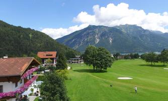 A picturesque landscape with a golf course, surrounded by mountains and green grass. In the foreground stands a charming building with blooming plants.