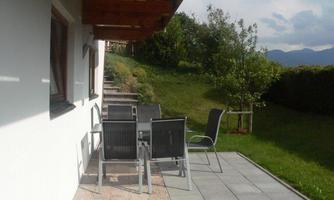 An outdoor area with a table and chairs on a paved surface. In the background, there is a green meadow and some trees.