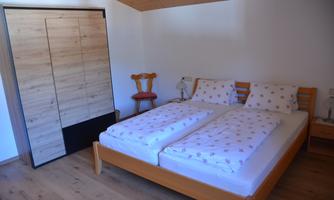 A cozy bedroom with a large double bed and bright wooden furniture. There is a wardrobe and a wooden chair on the wall.