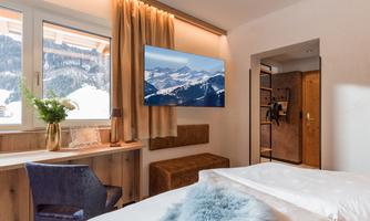A cozy bedroom with a large window through which the mountains are visible. There is a modern television and tasteful furniture.