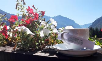 A cup of coffee on a table with colorful flowers in the foreground. In the background, mountains and a clear blue sky are visible.