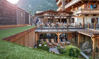 A modern restaurant with wooden architecture and a beautiful terrace. In the background, mountains and a green lawn can be seen.