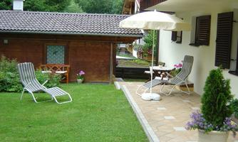 A beautiful garden with loungers and a table under a sunshade. In the background, you can see a wooden house and green trees.
