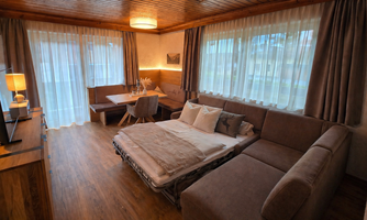 A cozy living room with a sofa bed and a log. There are large windows with curtains and a dining area with a table and chairs.