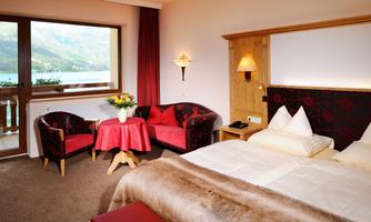 A cozy hotel room with a double bed, a table, and two armchairs. The view from the window shows a beautiful landscape with mountains and water.