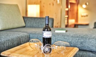 A red wine is placed on a wooden table, surrounded by overturned wine glasses. In the background, a cozy living area can be seen.