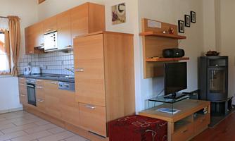 A modern kitchen made of wood with a dining area. In the background, there is a television and a cozy spot to relax.