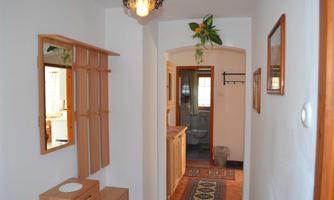 A bright hallway with a wooden wardrobe and a decorative lamp. To the right, a door leads to another room, while the floor is adorned with a carpet.