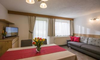 A bright living room with a cozy sofa and a dining table.  
Large windows let in plenty of light and a flower vase sits on the table.