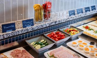 A buffet with various dishes, including ham, cucumbers, tomatoes, and eggs. The selection is attractively arranged and labeled.