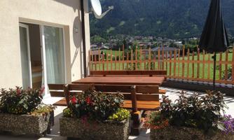 A terrace with a large wooden table and benches. In the background, mountains and a blue sky can be seen.