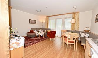A bright living room with a cozy sofa and a dining area. The room has large windows and a warm wooden floor.