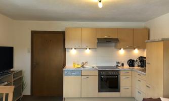 A modern kitchen with light wood cabinets and an island. The room is cozily furnished and features a television and a seating area.