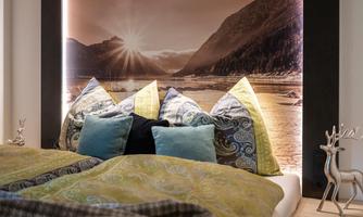 A cozy bedroom with a large bed, colorful cushions, and an impressive wall decoration. Soft light highlights the landscape in the background.