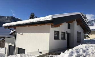 A modern house in the snow with a snow-covered roof. In the background, mountains and blue sky are visible.