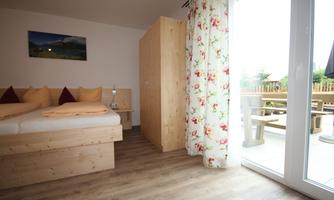 A cozy room with a wooden bed and flower-patterned curtains. Large windows let in plenty of light and lead to a terrace.