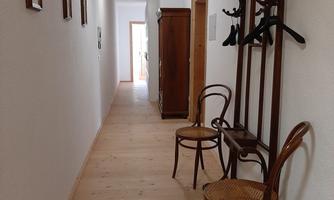 A bright hallway with wooden flooring and white walls. Pictures hang on the wall, and there are two chairs and a coat rack.