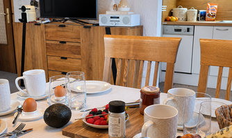 A laid breakfast table with various foods, including rolls, avocado, and fresh berries. In the background, a television and a modern kitchen are visible.