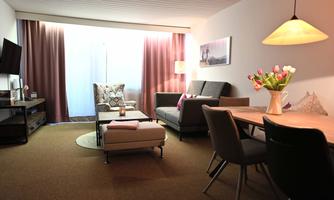 A modern living room with comfortable furniture and a large window. A dining table with a floral arrangement adds an inviting touch.