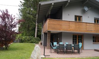 A modern house with a balcony and a terrace. The garden is green and well-maintained, with a table and chairs outdoors.