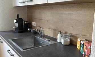 A modern kitchen with a dark work area and wood paneling. On the countertop are a coffee machine, sinks, and various food containers.