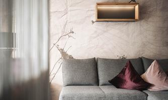 A modern living room with a gray couch and decorative pillows in pink and dark red. The wall is adorned with a subtle pattern and a wall shelf.