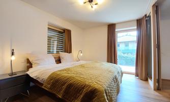 A modern bedroom with a large bed and a golden bedspread. Natural light falls into the room through the windows.