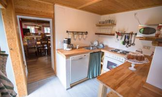 A modern kitchen with wooden shelves and bright walls. In the background, there is a dining area with wooden chairs and a table.