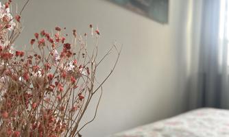 A cozy bedroom with a bed and floral bedding. In the foreground, there is a dried flower arrangement.