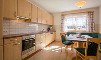 A modern kitchen with wooden cabinets and a dining table. The room is bright and cheerful with daylight streaming through the window.