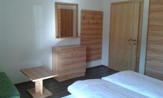 A simple and modern room with two beds, a wooden table, and a mirror. The walls are light-colored, and there is a wooden wardrobe.