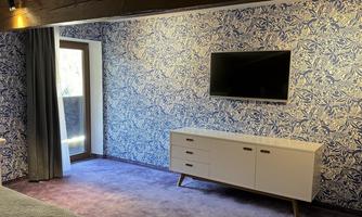 A modern designed room with blue wallpaper and a television on the wall. Below is a light chest of drawers and a window with curtains.