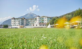 A modern hotel overlooking a mountain, surrounded by a green meadow. In the foreground, there are sun loungers and a pleasant atmosphere.