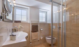 A modern bathroom with a glass shower and bright tiled floor. There is a sink, a towel rack, and windows that let in natural light.