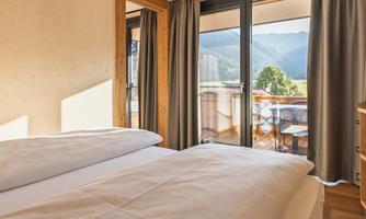A cozy bedroom with a large window that offers a view of the mountains. Outside, there is a balcony with seating.