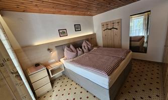 A cozy bedroom with a double bed and a wooden ceiling. The walls are light and there is a large window that lets in natural light.
