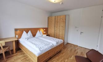 A bright room with a large wooden double bed. There is a table, a chair, and a wardrobe.
