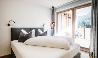 A modern bedroom with a large bed and elegant pillows. The room features a window front with access to a balcony and a view of nature.