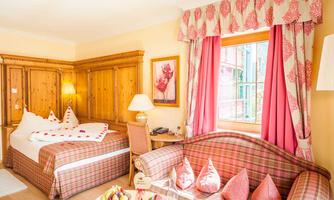 A cozy room with wooden paneling, a large bed, and a colorful sofa. The windows are adorned with red curtains, and there is a decorative selection of cushions.