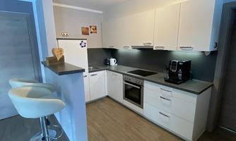 A modern kitchen with white furniture and wooden flooring. The kitchen is well equipped and has an appealing bar with stools.