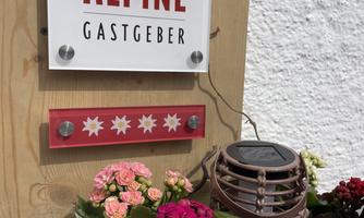 A sign with the inscription "ALPINE GASTGEF" on a wall. Below it, a flower box with colorful flowers.