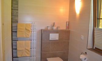 A modern bathroom with a WC, towels, and warm lighting. The walls are done in light colors, and there is a privacy screen at the window.
