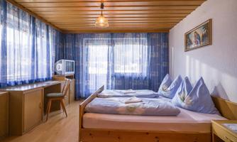 A cozy bedroom with two beds, wood paneling, and blue curtains. By the window, there is a small table with a chair and a television.
