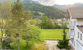 A view of green meadows and trees with mountains in the background. In the foreground, a residential house is visible.