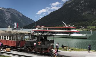 A picturesque landscape with a historic train and a large ship at the water. In the background, there are high mountains and a clear sky.